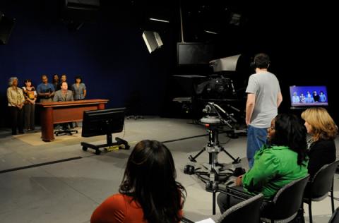 President Broderick is seated at a desk in the video studio, with a large video camera in front and employees from the Office of Distance Learning in the audience.