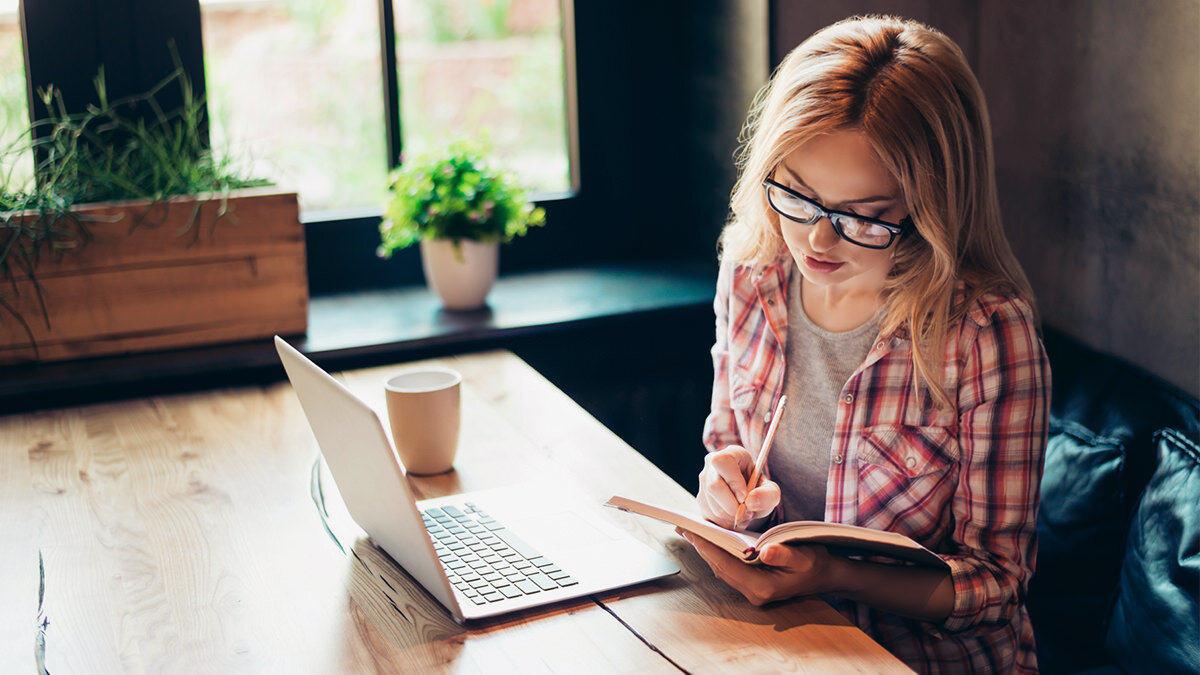 A woman studies in an online degree program while seated at a table with an open laptop and a notebook.