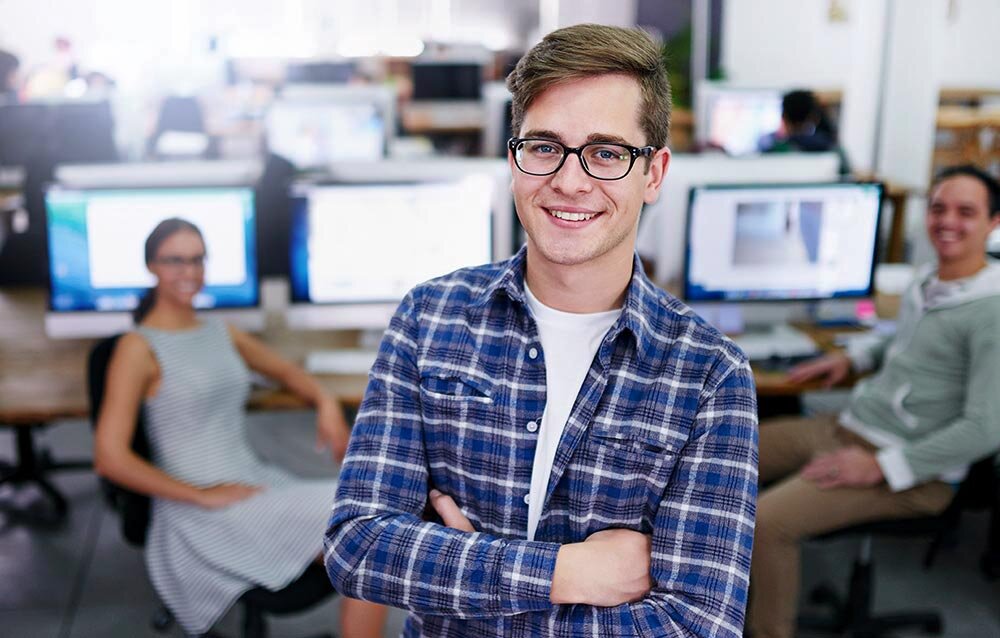 Young college student stands in an airy computer lab. His arms are folded and he is smiling at the camera.
