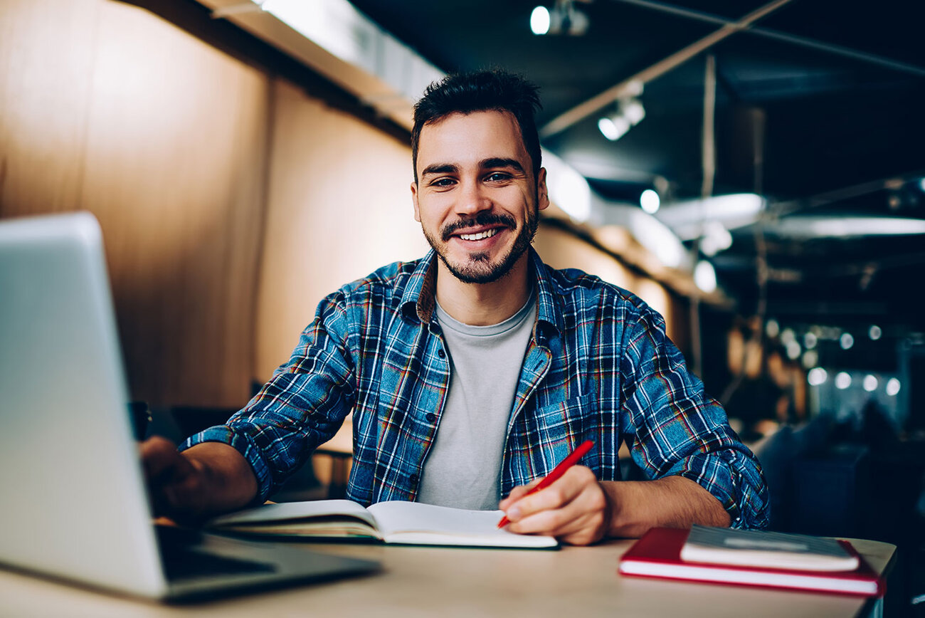 Smiling student sits at computer desk with a laptop and paper notebook