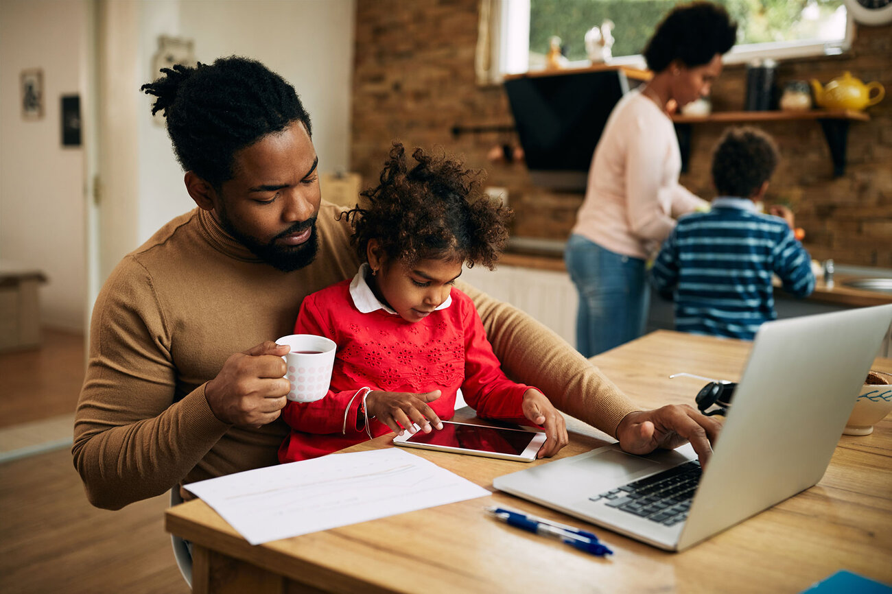 Dad works to transform his future with an online PhD program from ODUGlobal. While he sits working on his laptop his child looks at the screen and his family is in the background.