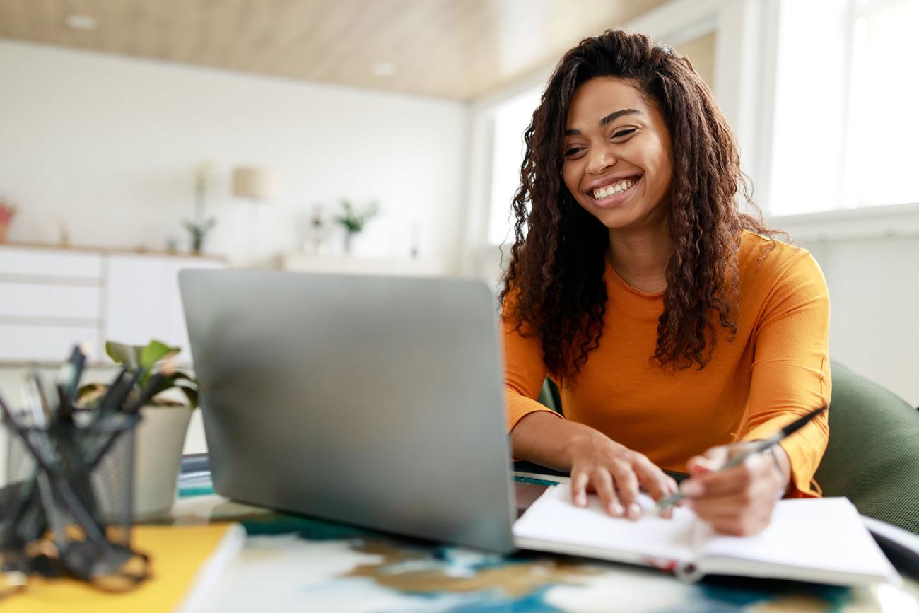 Woman in gold sweater sits at a desk and smiles at her computer while writing