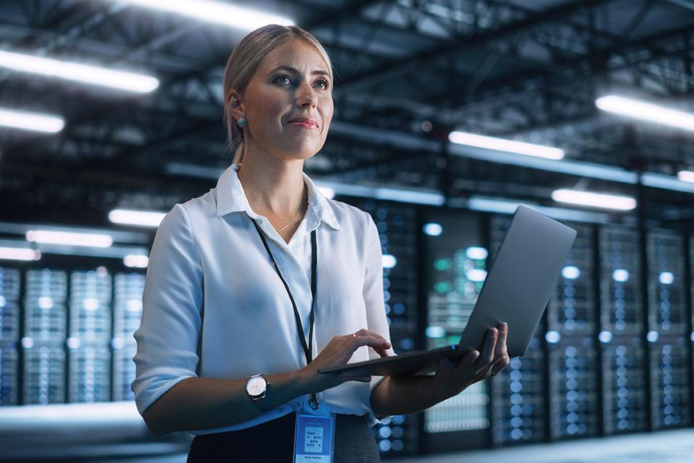 A data science professional stands holding a laptop, with server racks in the background