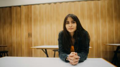 Woman sits at a desk in a classroom and looks at the camera intently 