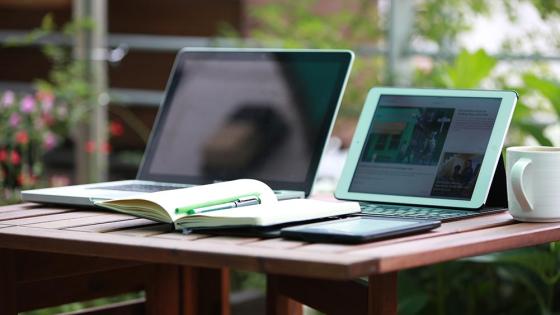 Two open laptops on a wooden desk with a paper notebook nearby