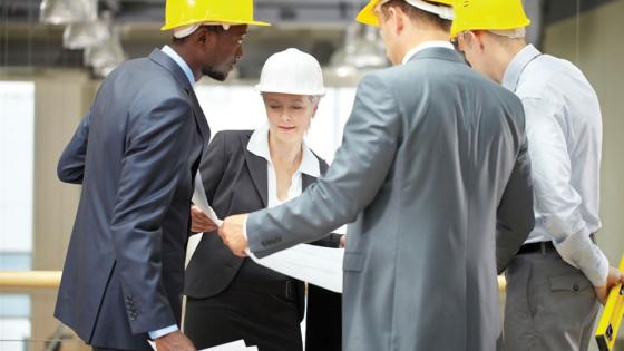 Engineering management staff gather around a large print out in a warehouse setting.