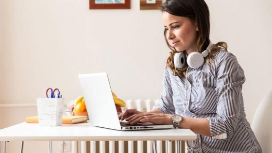 Woman with headphones around her neck types on a laptop seated at a kitchen table.