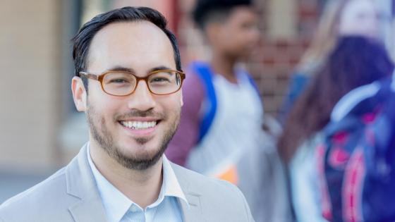 Man wearing a button shirt and sport coat smiles at the camera in a school hallway.