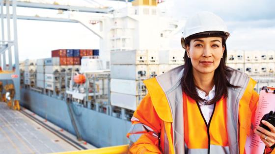 A woman in a hard hat and orange safety jacket stands on a balcony overlooking a cargo port where containers are being unloaded from a shipping vessel.