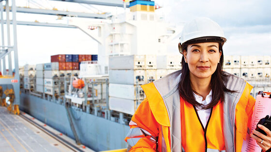 A woman in a hard hat and orange safety jacket stands on a balcony overlooking a cargo port where containers are being unloaded from a shipping vessel.