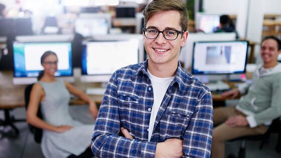 Young college student stands in an airy computer lab. His arms are folded and he is smiling at the camera.