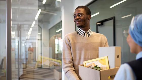 A smiling, young African American man arrives at his new job holding a box of items for his new office.