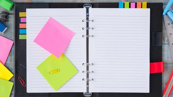 Overhead view of an open notebook with sticky notes, pens, and paperclips scattered around it on the table.