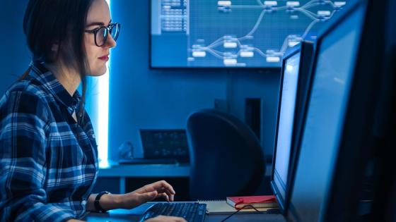 A female professional in the cybersecurity field works at a computer on a desk with dual monitors.