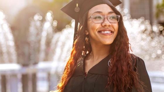 An ODU graduate wearing a graduation cap and gown smiles in the sunlight near ODU's lion fountain.