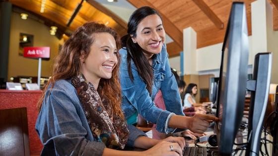 A female librarian guides a young student to find information at a desktop computer.