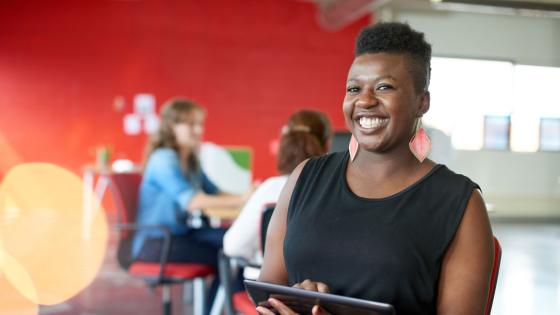 A smiling woman in an open office setting works on a tablet device.