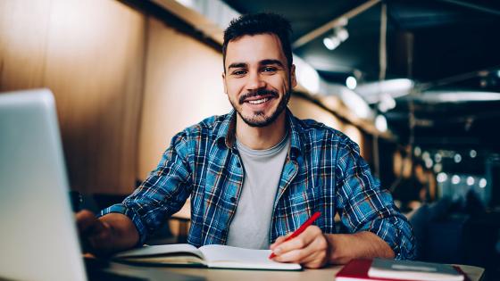 Smiling student sits at computer desk with a laptop and paper notebook