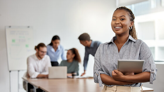 Woman with tablet in a boardroom with colleagues meeting behind her 