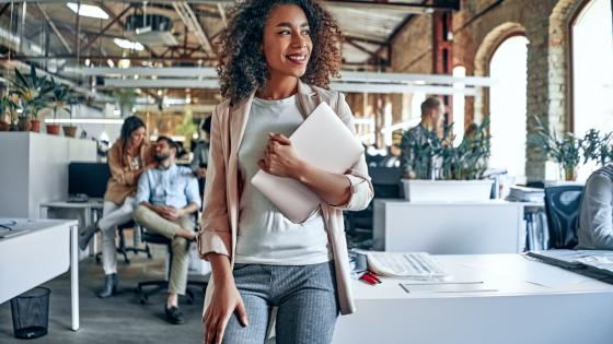 Woman stands in a professional workspace holding a laptop and smiling