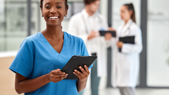 A female nurse wearing a blue uniform in a hospital corridor pauses to smile at the camera.