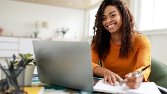 Woman in gold sweater sits at a desk and smiles at her computer while writing