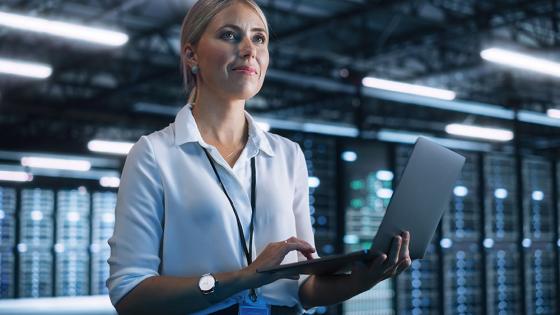 A data science professional stands holding a laptop, with server racks in the background