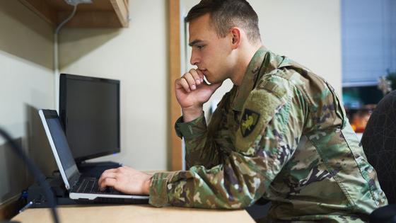 An active duty servicemember prepares for class at a laptop on a desk.