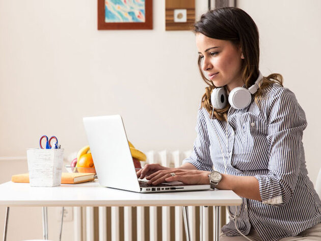 Woman with headphones around her neck types on a laptop seated at a kitchen table.