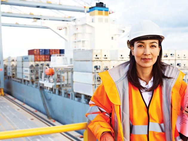 A woman in a hard hat and orange safety jacket stands on a balcony overlooking a cargo port where containers are being unloaded from a shipping vessel.