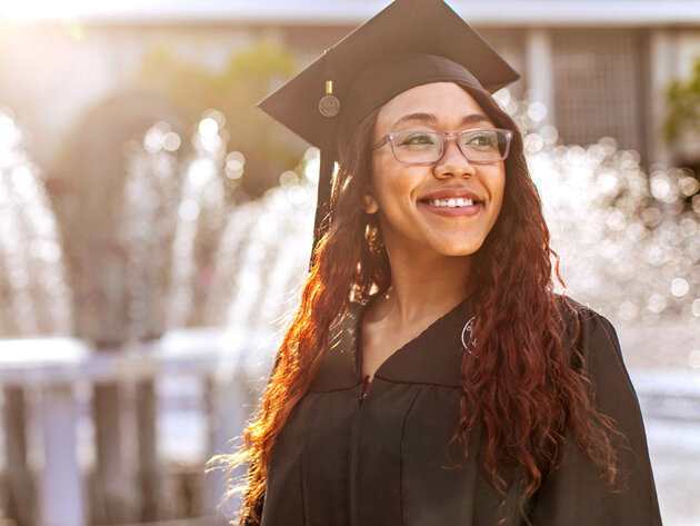 An ODU graduate wearing a graduation cap and gown smiles in the sunlight near ODU's lion fountain.