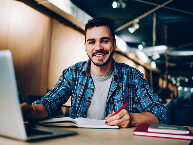 Smiling student sits at computer desk with a laptop and paper notebook