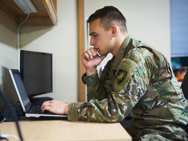 An active duty servicemember prepares for class at a laptop on a desk.