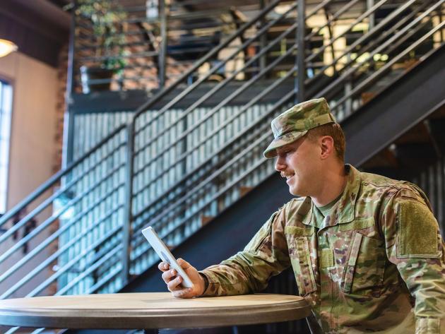 A seated active duty military service man looks at a handheld tablet device.