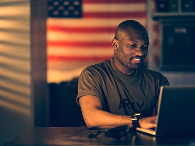 Military service member sits at a computer in front of the American flag