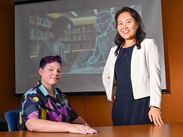 Dr. Jinhee Kim and PhD candidate Rita Detrick pose for a portrait in front of a screen with a presentation about AI.