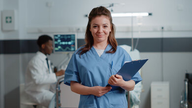 Smiling woman wearing scrubs stands with a clipboard in a room with medical equipment in the background